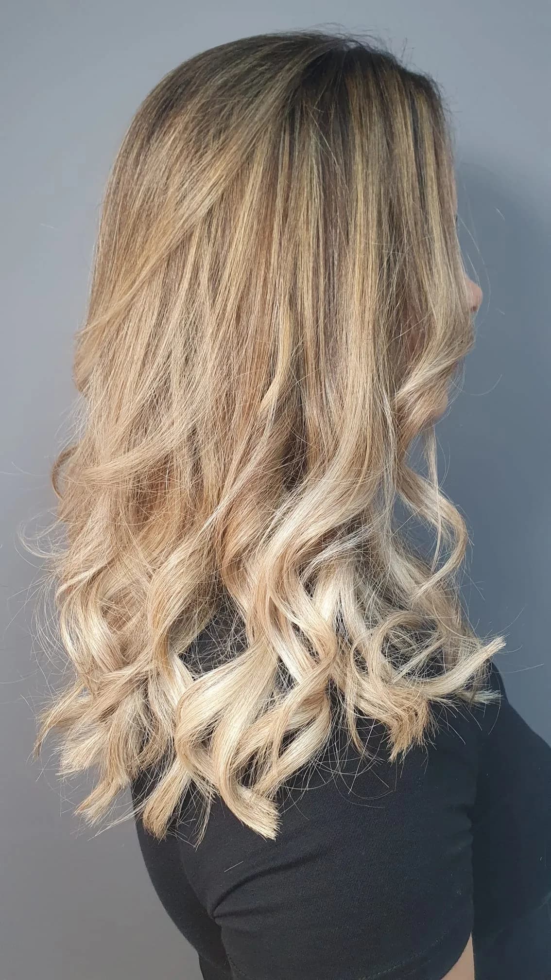 Longs cheveux blonds vus de trois quarts dos, racines foncées et larges boucles éclaircies vers les pointes