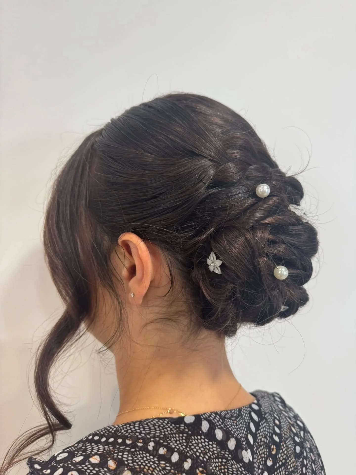 Chignon bas tressé sur cheveux bruns, vu de trois quarts dos, orné de perles et d'une petite fleur blanche